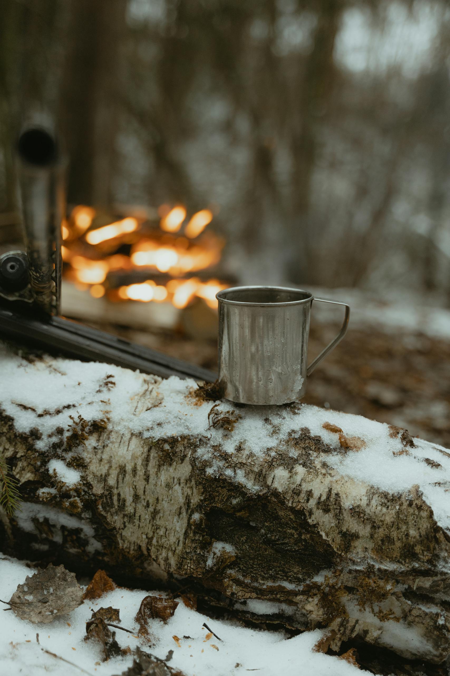 Metal cup and firewood by a winter campfire on snow-covered forest log.