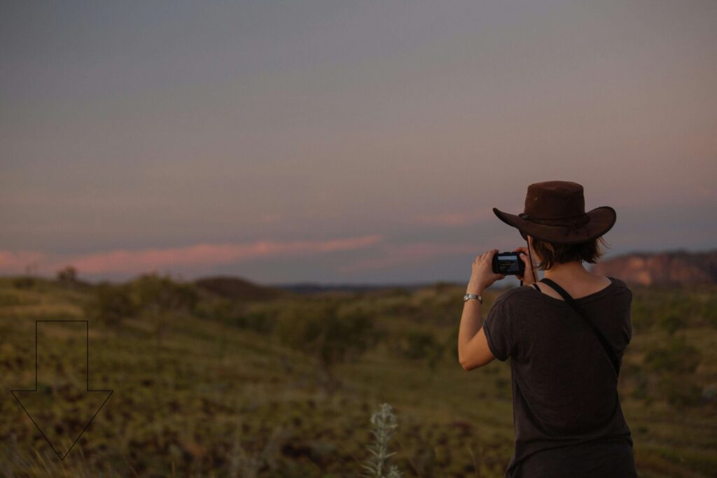 A person wearing a brown hat captures a scenic sunset over a vast outdoor landscape using a smartphone device while camping in nature.